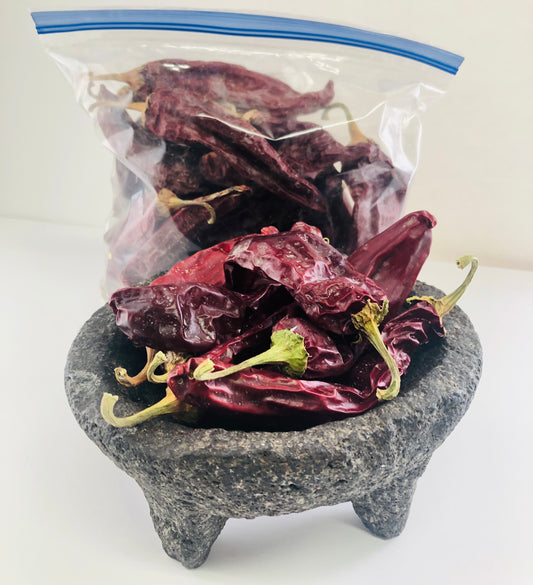 A clear plastic bag filled with dried red chile peppers (pods) placed on top of a traditional Mexican molcajete.